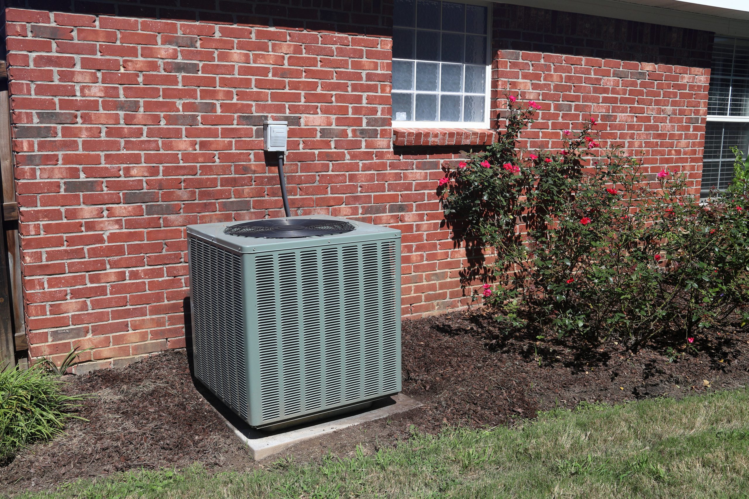 Air Conditioner system next to a home, with rose bushes and brick wall.